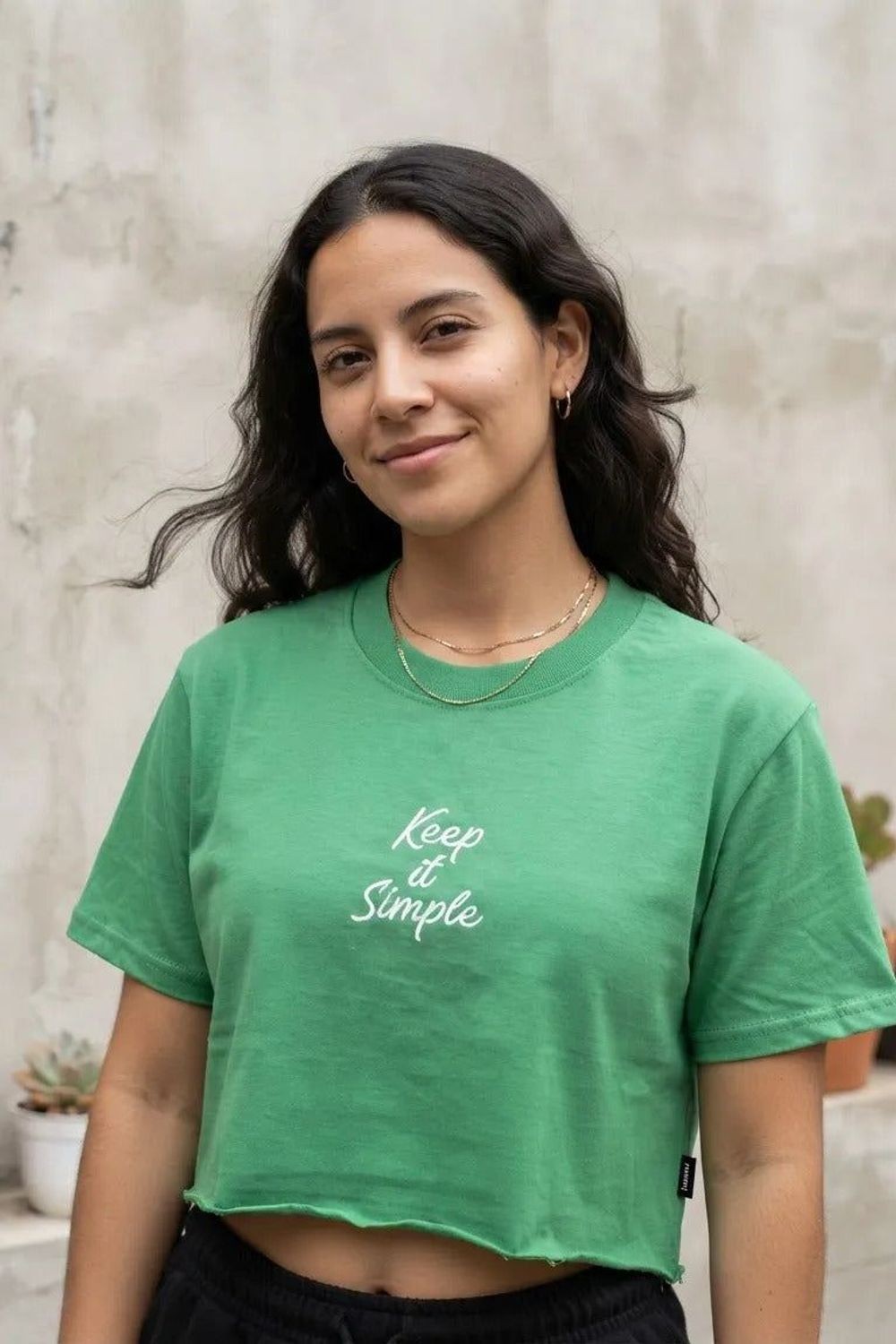 Blusa cropped Lerrux Keep Is Simple verde, vestida em modelo sorridente com cabelo cacheado em frente a parede de concreto Imagem principal do produto