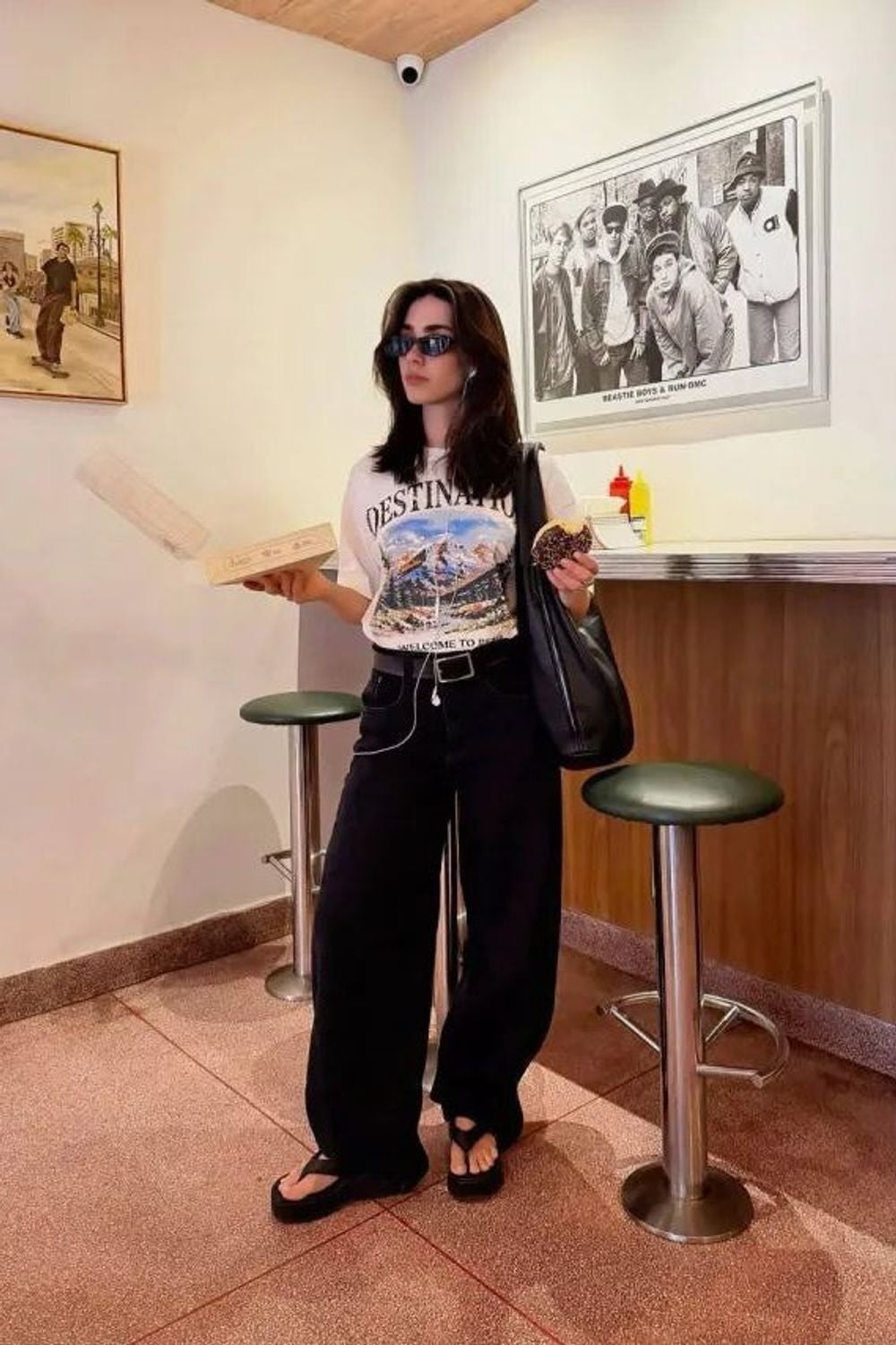 Calça Balloon Relaxed Lerrux preta, com modelo de camiseta branca e bolsa de ombro, em frente a um balcão de lanchonete
