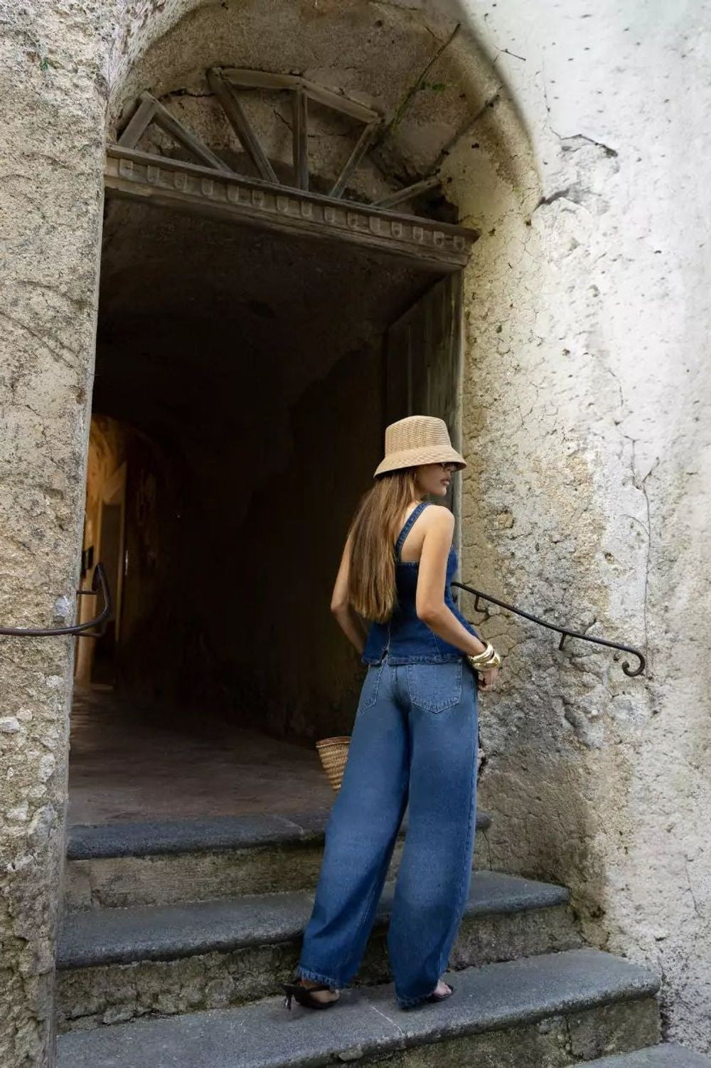 Calça jeans balloon Alcance Dolce Vita azul, vestida na modelo de costas, em escadaria de pedra com chapéu de palha Imagem principal do produto