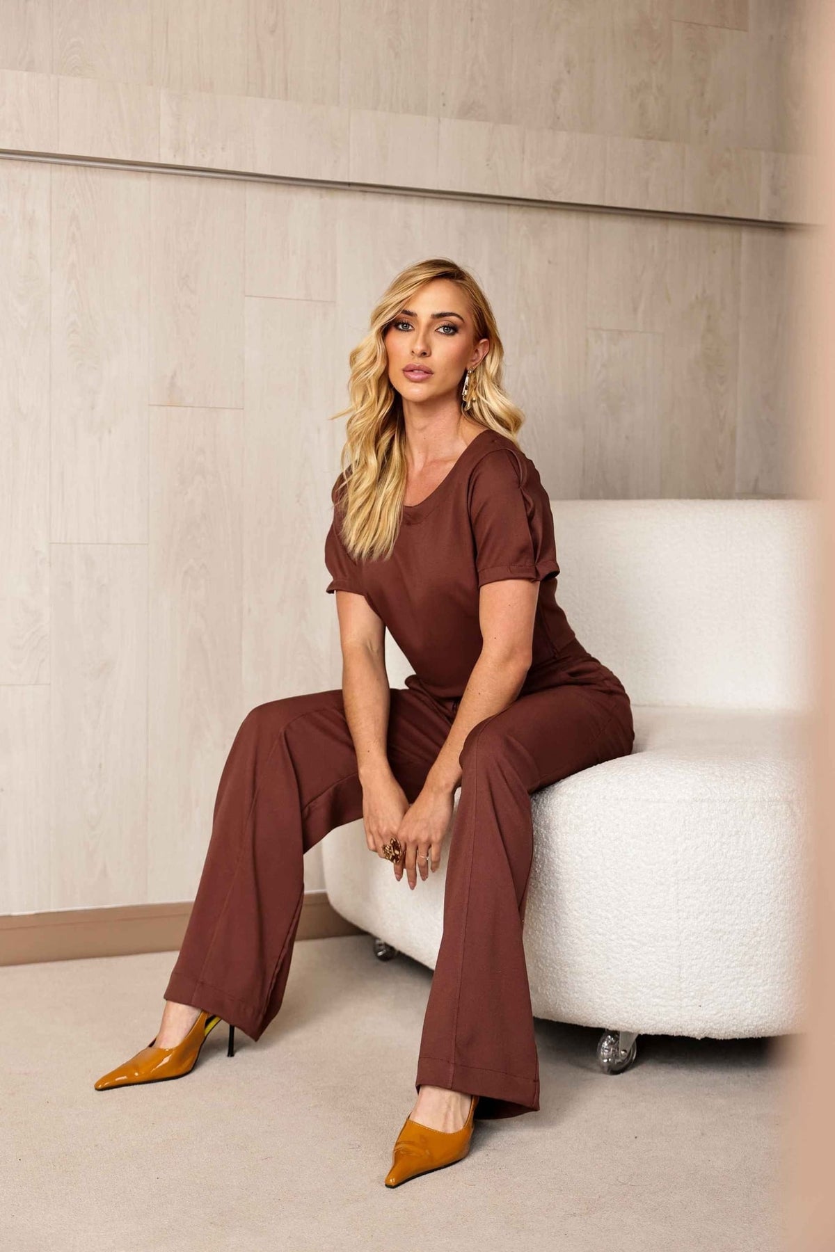 Modelo sentada em sofá branco usando conjunto Amicia marrom, com calça pantalona e sapato amarelo de salto