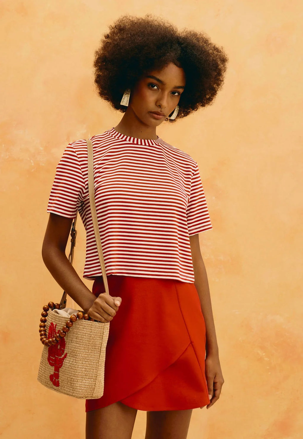 Camiseta Cacay cropped de listras vermelhas e brancas, vestida na modelo com saia vermelha e bolsa de palha